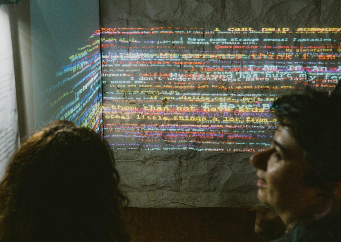 Two visitors stand in front of a corner-projected wall of overlapping multicoloured confessions